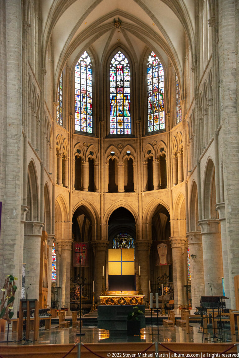 Closer centered view toward the cathedral sanctuary, with three stained-glass windows, ribbed vaulting, and pale stone arcades.