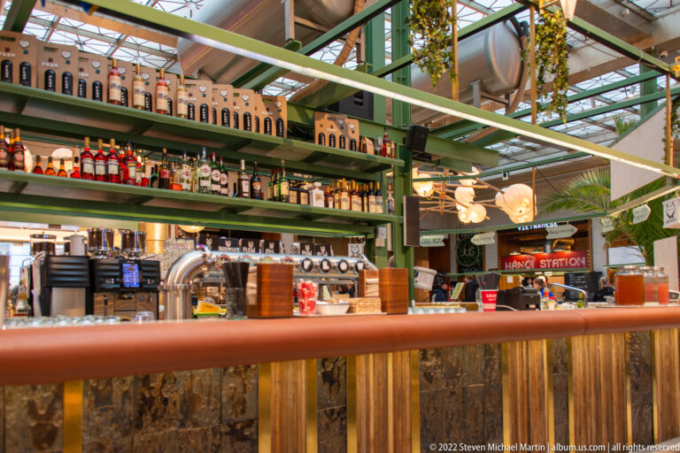 Food-hall bar with bottle shelves, beer taps, green steel framing, hanging plants, and mixed industrial finishes.