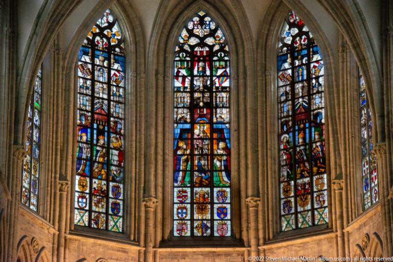 Tall Gothic stained-glass windows with heraldic shields, standing figures, and dense red, blue, gold, and white panels.