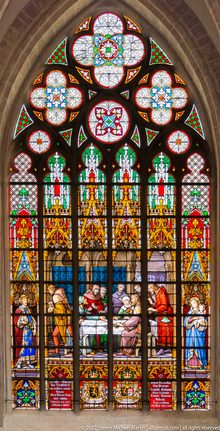 Five-lancet stained-glass scene with a reclining person on a bed or bier, surrounded by attendants beneath gold canopies.