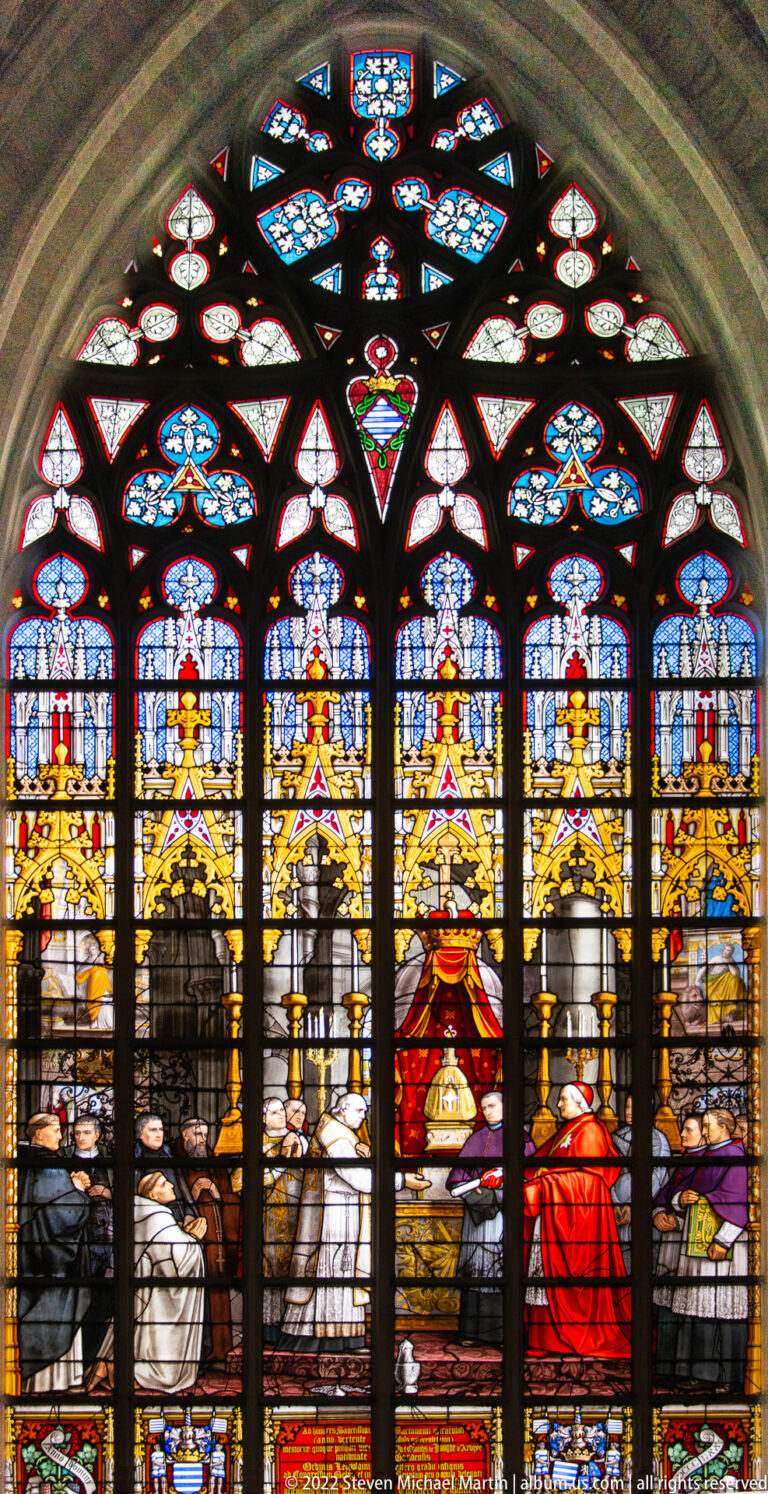 Arched stained-glass window with a seated central figure, robed attendants, a red-clad participant, and dense upper tracery.