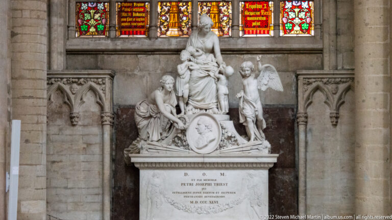 White marble funerary monument with a seated central female figure, attendants, shield, and inscribed pedestal beneath stained glass.