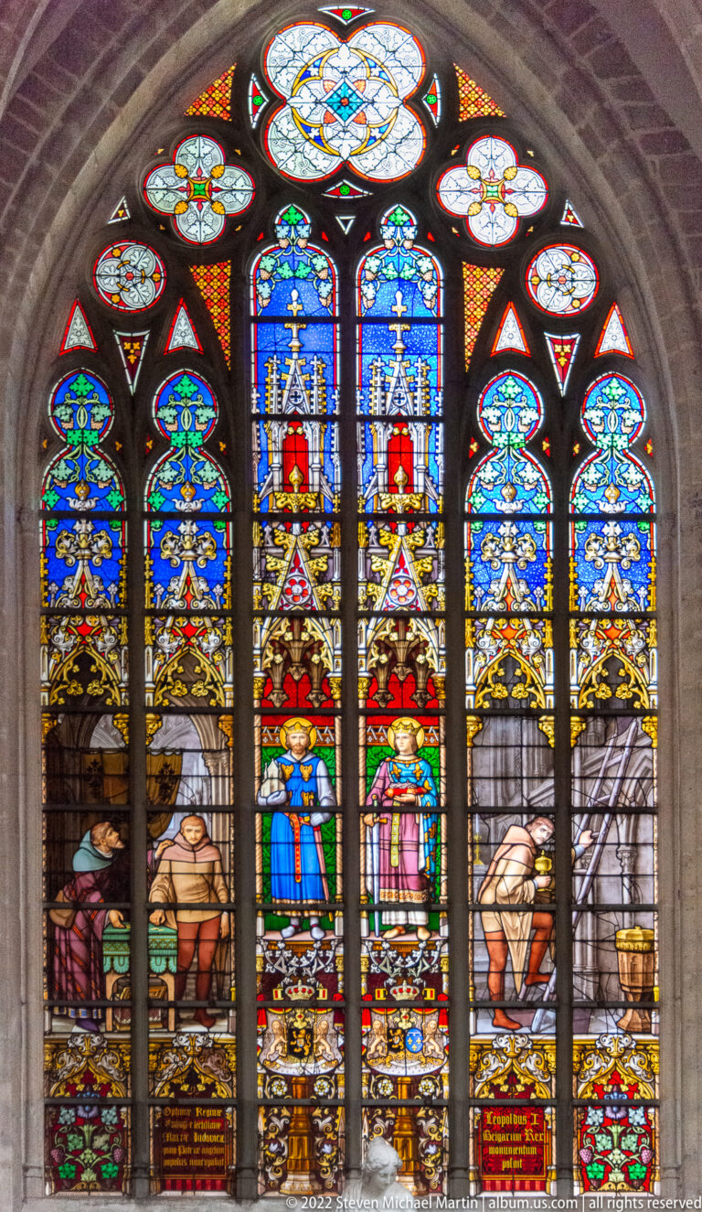 Arched stained-glass window with a central standing pair, side narrative scenes, blue-and-gold canopies, and a large rosette above.