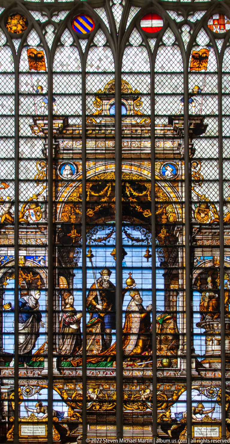 Close detail of a narrative stained-glass scene in brown, gold, white, and blue, seen through a protective mesh.