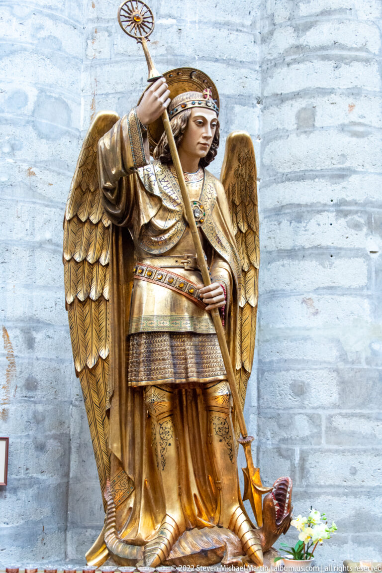 Gilded winged angel in patterned robes holding a long staff against a pale stone wall, with flowers at its feet.