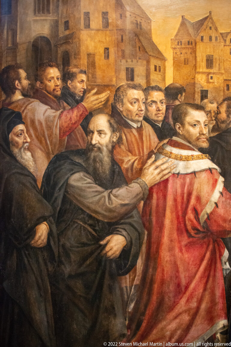 Detail of a religious painting showing a bearded man in dark robes beside clerics and townspeople in an urban setting.