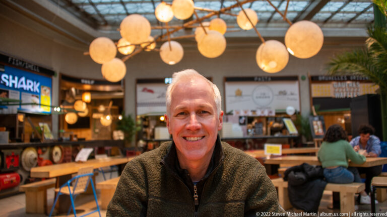 Smiling man seated at a communal table in a Brussels food hall beneath globe lights, with vendor counters behind him.