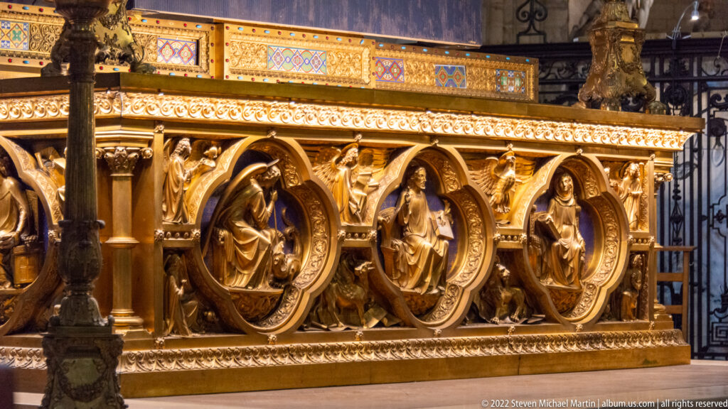 Gilded church furnishing with sculpted standing figures in round arches, surrounded by carved foliage and colorful decorative bands.