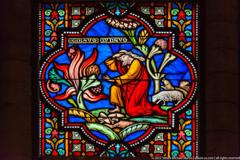Stained-glass medallion with a robed biblical figure beside flames and a pale animal, surrounded by colorful floral ornament.