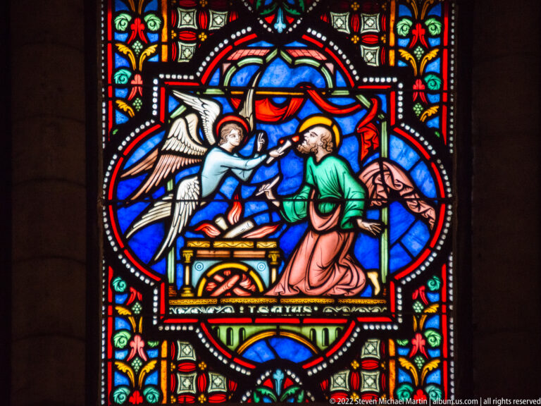 Stained-glass scene of an angel reaching toward a bearded man beside a small altar-like structure on a blue background.