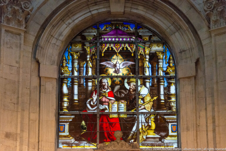 Arched stained-glass church window with central sacred figures, ornate canopy work, and heraldic shields in a pale stone wall.