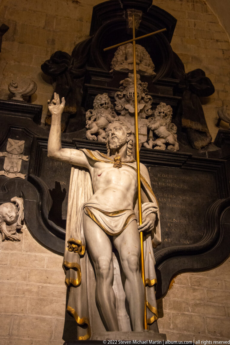 Standing pale stone figure of Christ with raised hand and long staff, set before a dark sculpted memorial.