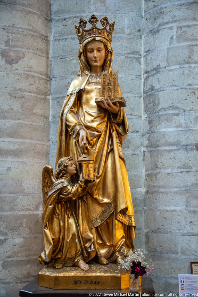 Crowned gilded Virgin holding the Christ Child, with a smaller kneeling figure beside her against a pale stone wall.