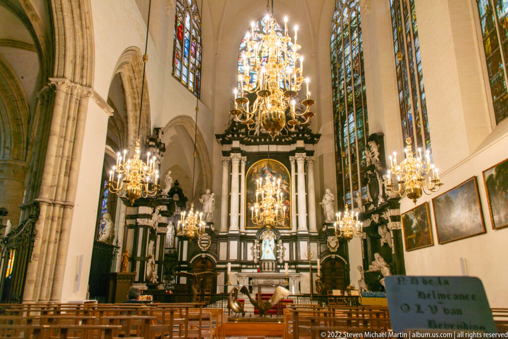 Church interior with rows of chairs, multiple crystal chandeliers, stained-glass windows, pointed arches, and a dark altar at the far end.