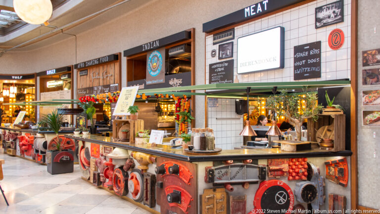 Decorated indoor food stall with menu boards, serving counter, produce, lamps, and colorful vintage objects covering the frontage.