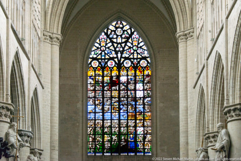 Tall pointed stained-glass window filled with many small scenes and shields, framed by pale Gothic stone piers.
