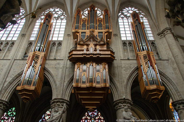 Large central pipe organ with two tall flanking pipe cases mounted against pale Gothic arches and windows.