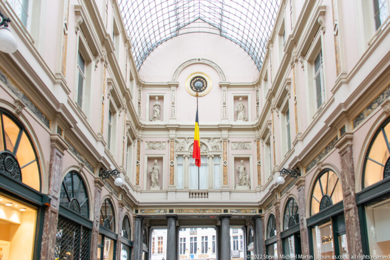 End of the Galeries Royales Saint-Hubert with Belgian flag, round clock, statuary niches, and the glass roof curving overhead.