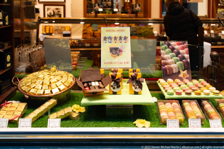 Shop window filled with boxed chocolates, pralines, and rows of pastel macarons on tiered display shelves.