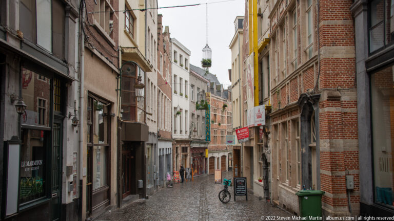 Narrow cobbled Brussels street lined with brick and stucco buildings, shop signs, bicycles, and a few pedestrians.