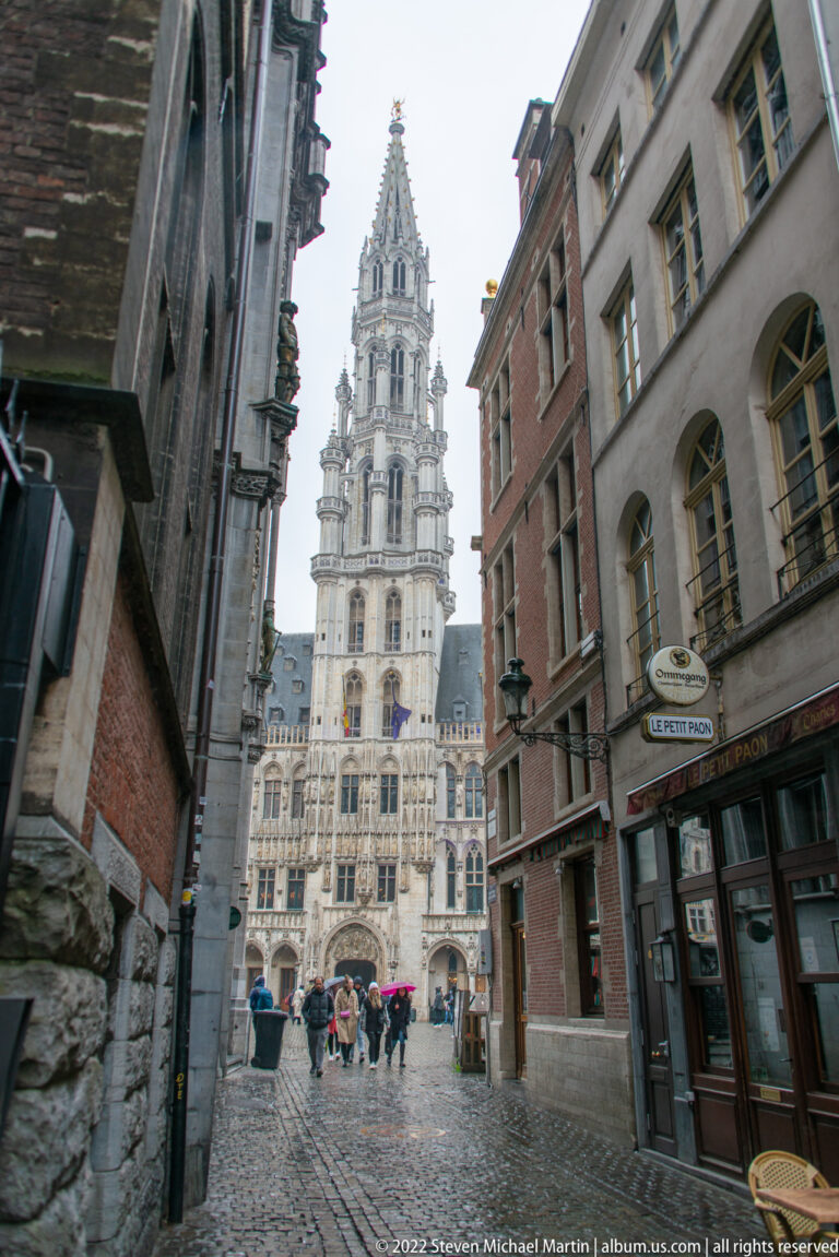 Brussels Town Hall tower seen at the end of a narrow cobbled street, framed by historic buildings on both sides.