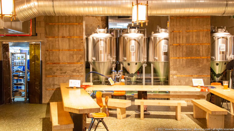 Stainless-steel brewery tanks behind wooden tables and benches in an industrial Brussels food-hall interior.