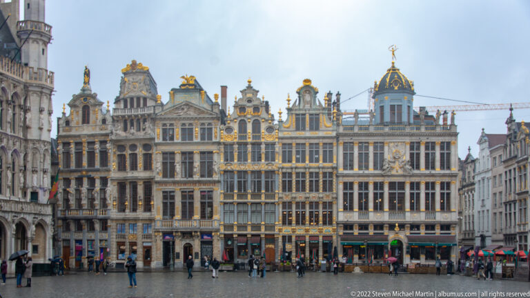 Row of ornate Brussels Grand-Place guildhouses with gilded trim, sculpted gables, many windows, shops, and pedestrians below.