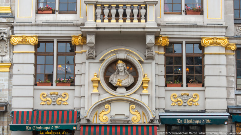 Close view of La Chaloupe d'Or façade with portrait bust in a round niche, gilded scrollwork, windows, and striped awning.