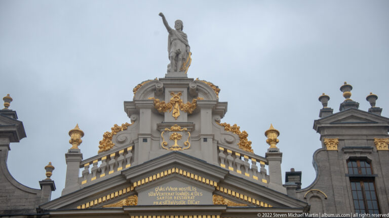 Ornate Grand-Place gable topped by a standing statue, with gilded scrollwork, urns, a cartouche, and Latin inscription below.
