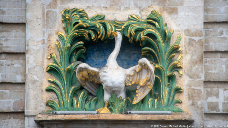 White swan relief with raised wings framed by green-and-gold foliage on a pale stone façade.