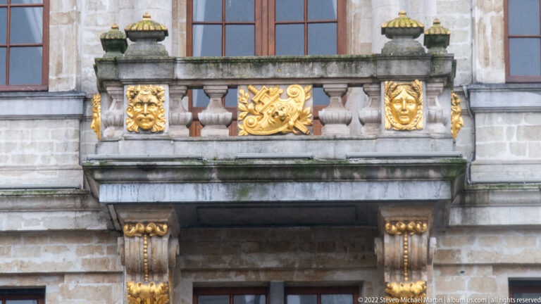 Stone balcony with gilded masks, central emblem, carved balusters, and gold-highlighted capitals beneath tall windows.