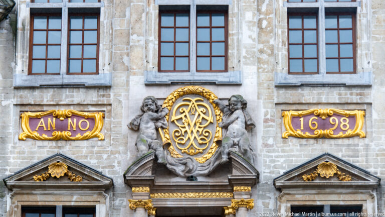 Grand-Place façade detail with gilded monogram, two putti, dark windows, and the date 1698 in gold.
