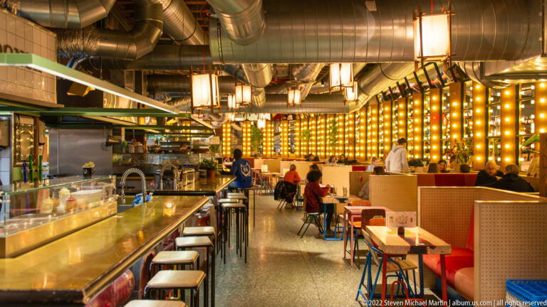 Long restaurant interior with open kitchen counter, stools, booths, exposed ductwork, and a glowing grid of square lights.