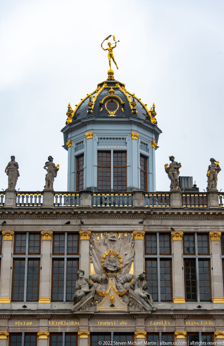 Upper façade of Le Roi d’Espagne with polygonal domed roof, gilded rooftop figure, sculpted busts, and gold trim.