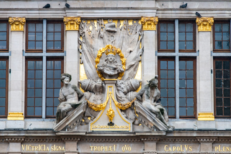 Le Roi d’Espagne façade with central portrait bust, draped stone surround, seated figures, gilding, and Dutch inscription below.