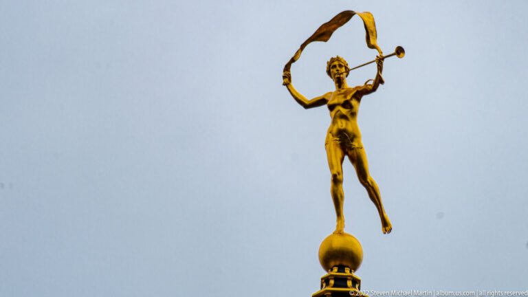 Gilded rooftop figure of Fame standing on a sphere, holding a flowing banner and trumpet against a gray sky.