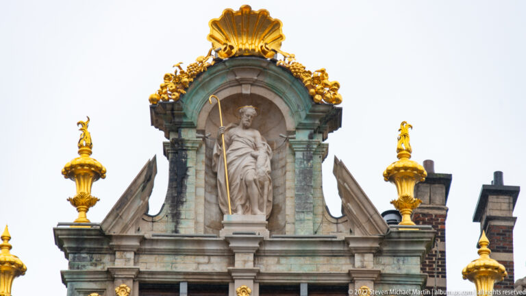 Stone standing figure in an arched niche beneath a gilded shell, with gold urns and ornament on the surrounding façade.