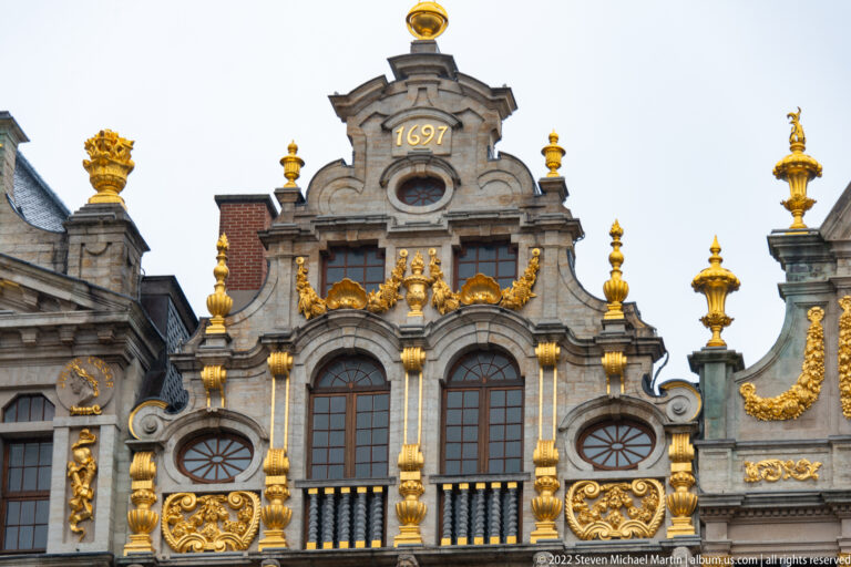 Upper façade of Le Roi d’Espagne with the date 1697, gilded urns, scrollwork, paired windows, and sculptural ornament.