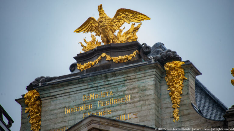 Gilded eagle with spread wings above a Latin inscription, garlands, and dark sculptural forms on a stone façade.
