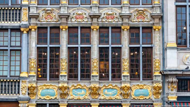 Le Roi d’Espagne façade with tall windows, gilded pilasters, carved masks, and blue-green inscription cartouches below.