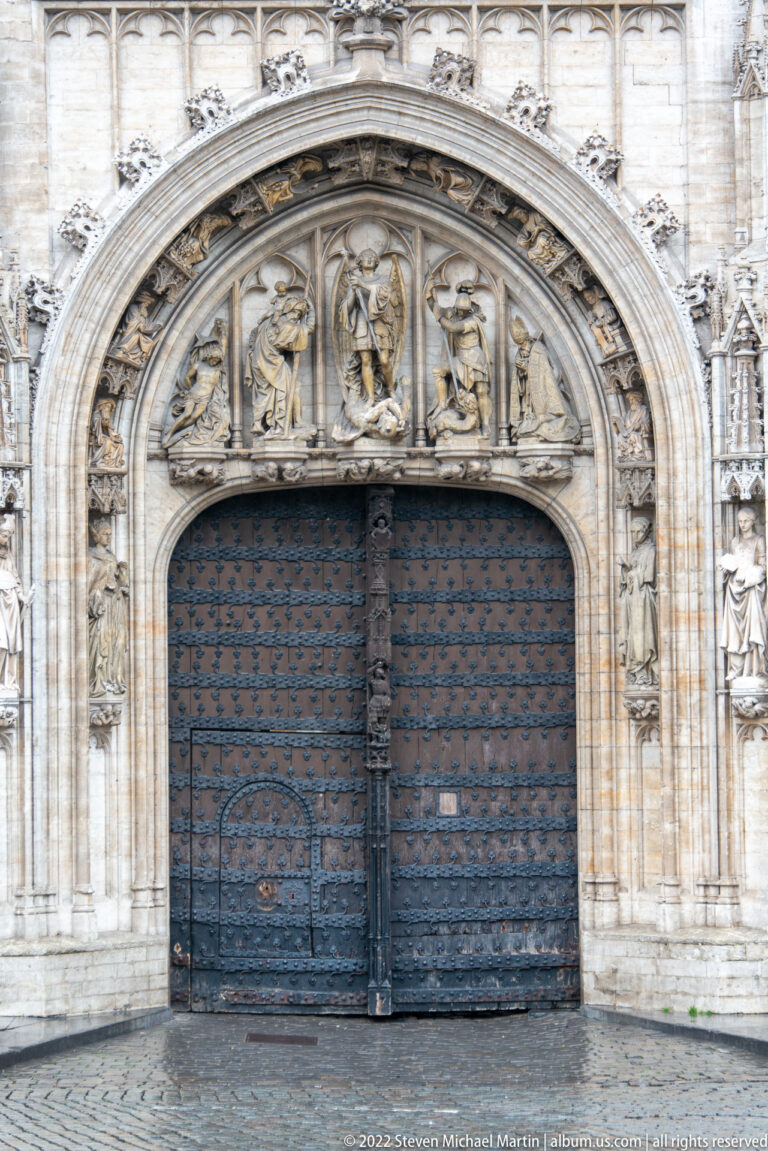 Large dark wooden doors beneath a richly carved Gothic arch with figures, foliage, tracery, and relief panels.