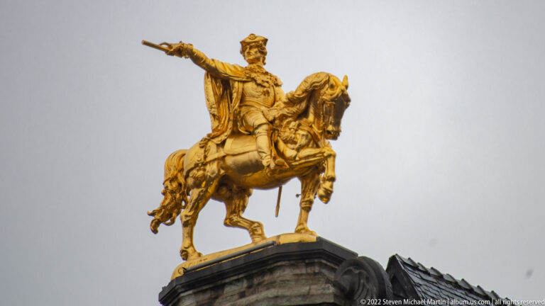 Gilded equestrian rooftop statue of a mounted ruler extending one arm, silhouetted against a pale gray sky.