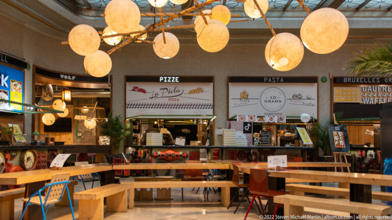 Communal tables and globe lights face pizza, pasta, waffle, and other vendor counters inside a Brussels food hall.