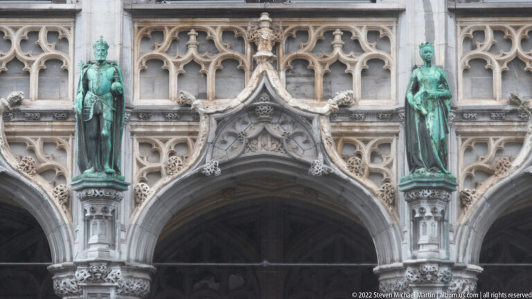 Two green-patinated standing statues flank a pointed Gothic arch surrounded by intricate pale-stone tracery.