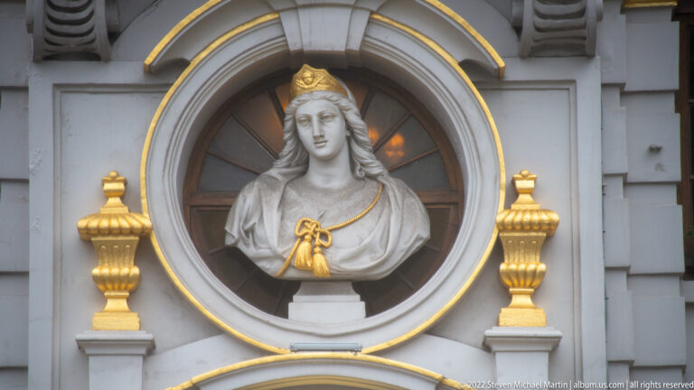 Crowned female stone bust in a circular niche with gold edging and two gilded finials below.