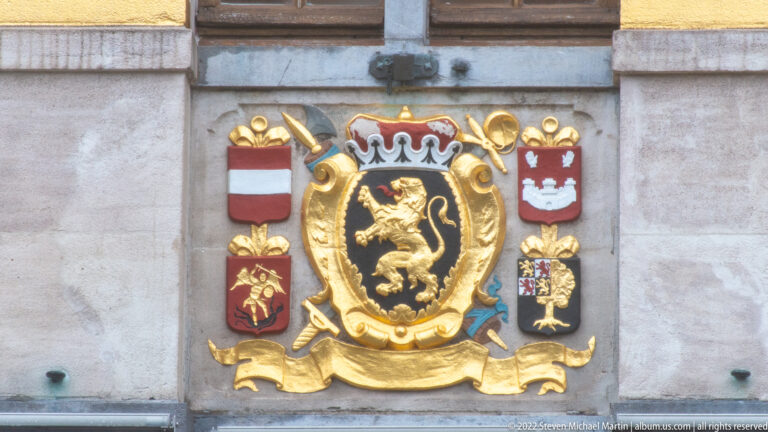 Large gilded lion coat of arms beneath a crown, surrounded by smaller shields and trophy-like gold ornament on stone.