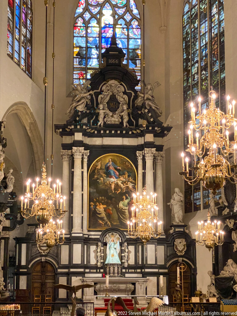 Baroque altar with large religious painting, pale columns, glowing chandeliers, and stained glass inside Brussels Cathedral.
