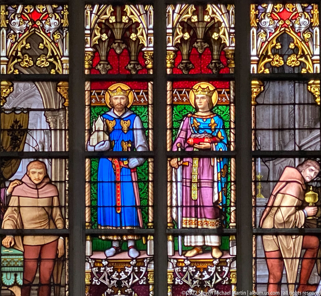 Stained glass with a crowned man and woman standing centrally, flanked by kneeling figures beneath Gothic canopy work.