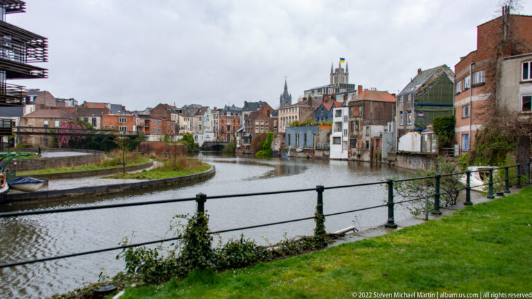 Curving Ghent canal bordered by historic houses, grassy banks, railings, and distant church towers under an overcast sky.