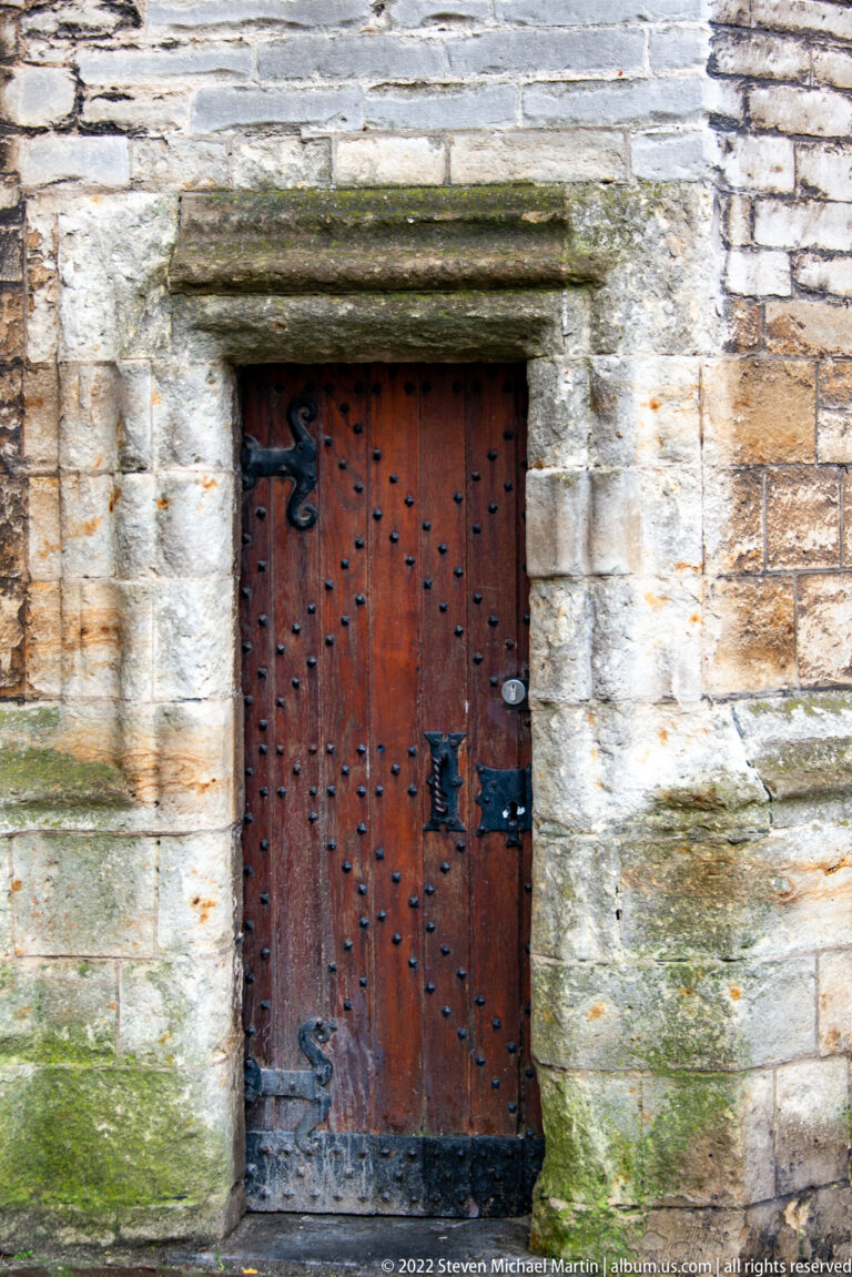Small reddish-brown studded wooden door with black iron hardware set in weathered pale stone.